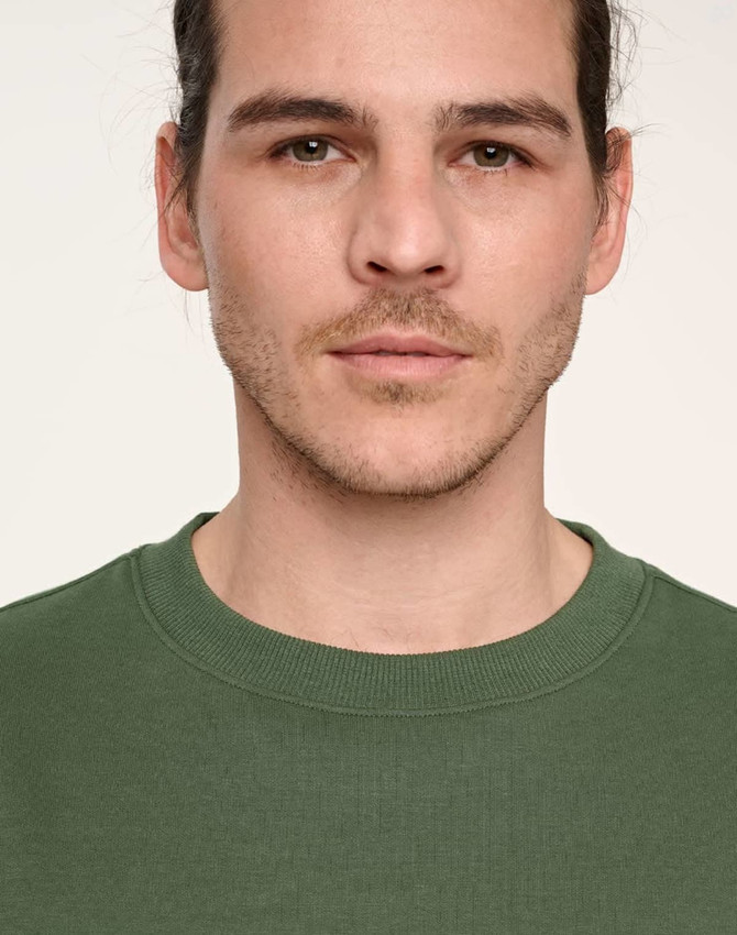 A close-up of a person wearing a green unisex crew neck sweater with a logo on the chest.