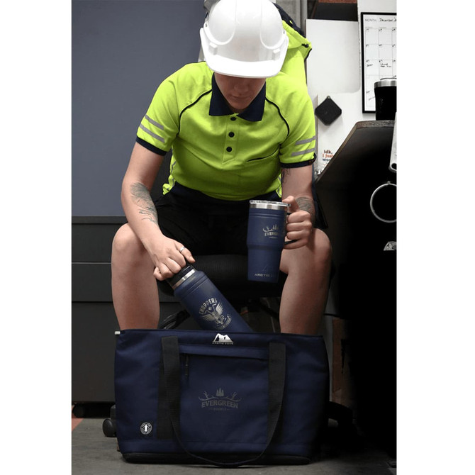 A blue drink bottle and mug sit next to a dark blue duffel bag, with a person in a high-visibility shirt and helmet.