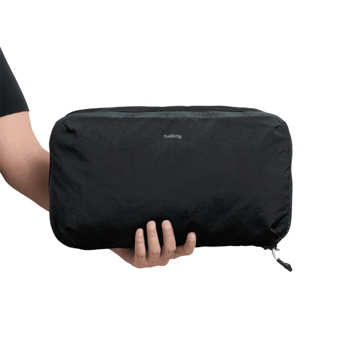 A black packing cube being held by a person, featuring a zippered closure and a small logo on the front.