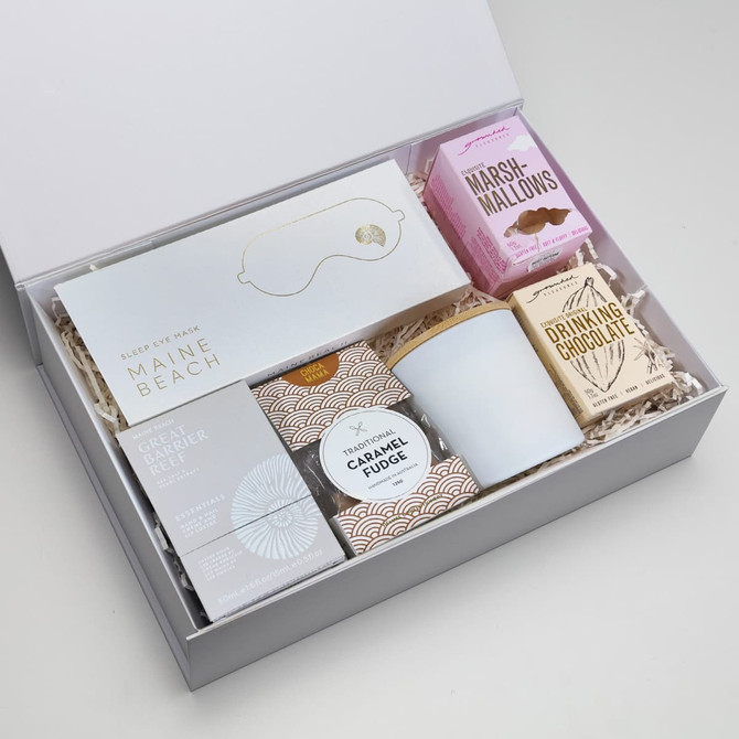 A white gift box contains a sleep mask, caramel fudge, drinking chocolate, marshmallows, and a candle, arranged with decorative filler.