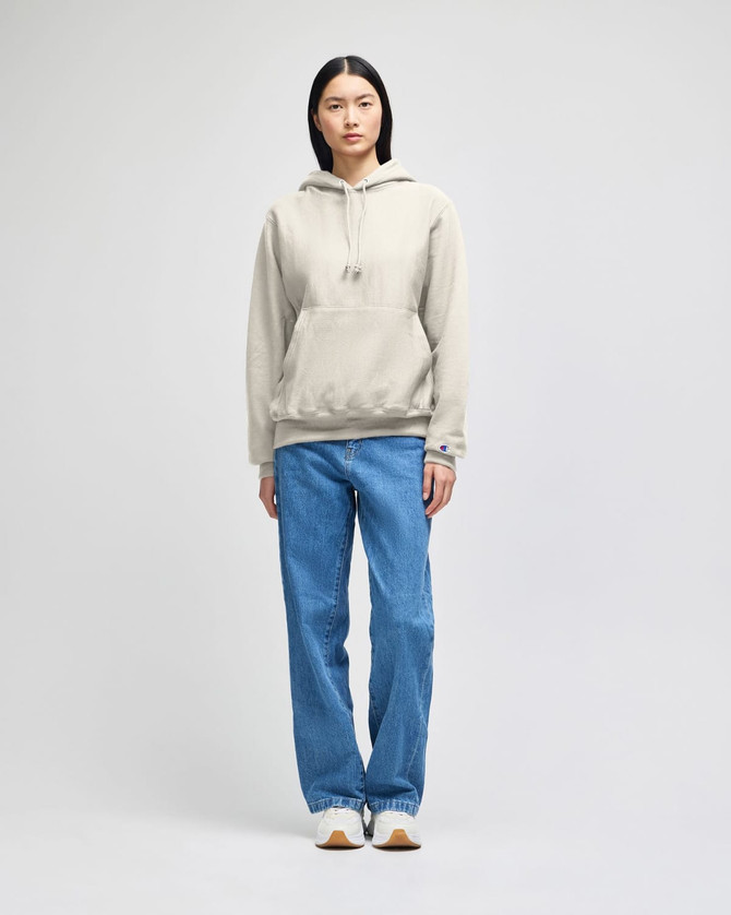 A model wears a beige Champion Reverse Weave® hooded sweatshirt paired with blue jeans. The sweatshirt features a logo.