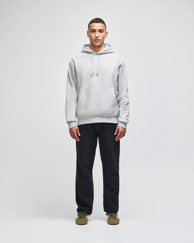 A man wearing a light grey hoodie with a hood, paired with dark pants, standing against a plain background.
