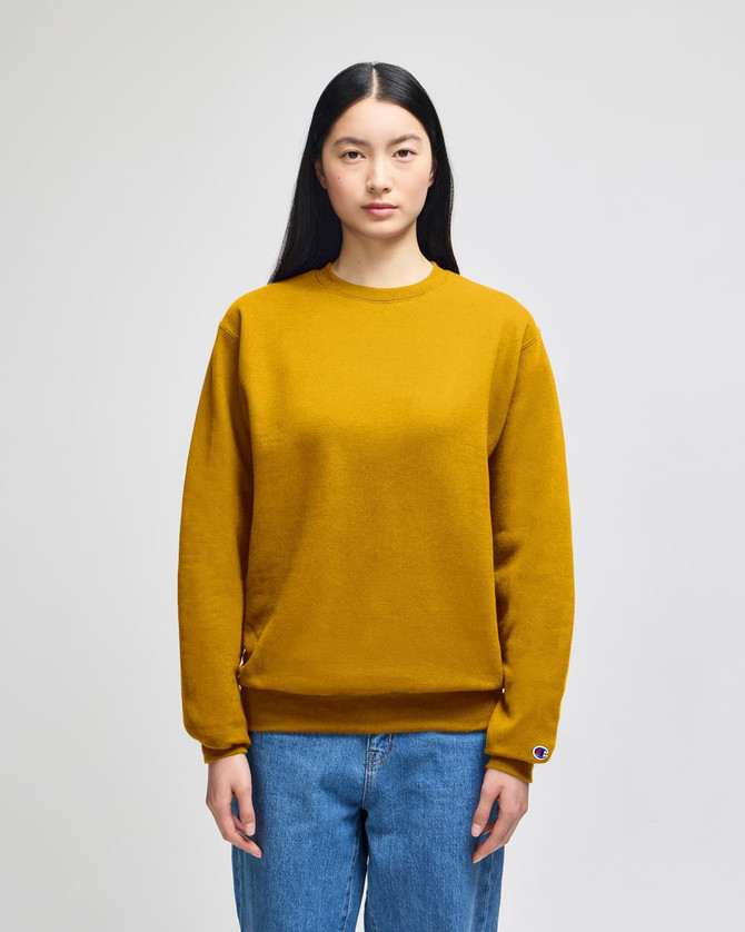 A mustard yellow adult crewneck sweatshirt with long sleeves, featured on a model wearing jeans. The sweatshirt has a logo on one sleeve.
