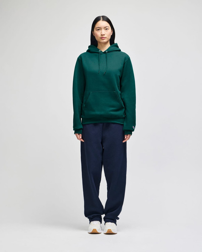 A green hooded sweatshirt with a front pocket, paired with navy trousers, displayed on a model.