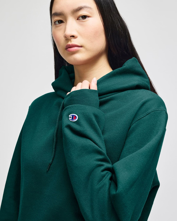 A model wearing a dark green Champion Powerblend® adult pullover hooded sweatshirt with a logo on the sleeve.