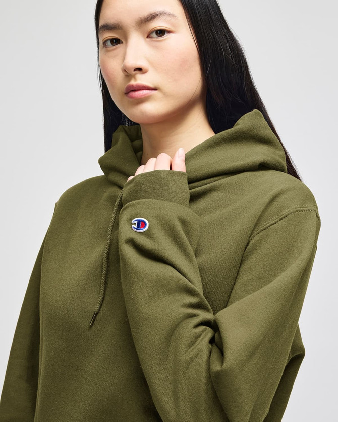 A woman models an olive green hooded sweatshirt with a logo on the sleeve.