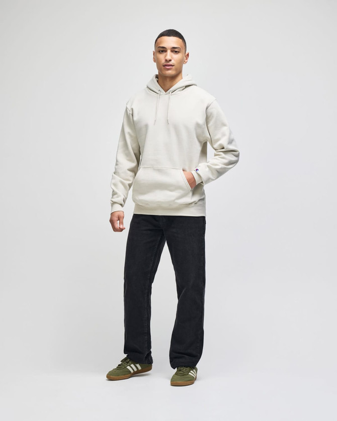 A light grey pullover hooded sweatshirt worn by a model, paired with dark trousers and shoes. The sweatshirt has a logo.