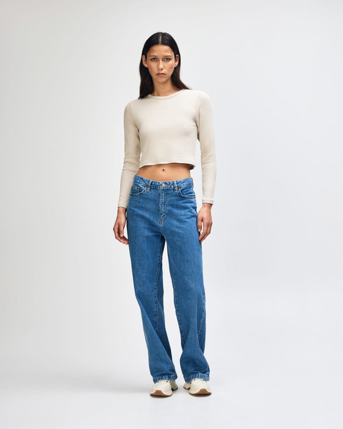 A fitted, long-sleeve, cropped t-shirt in beige, paired with blue jeans. The model stands on a plain background.