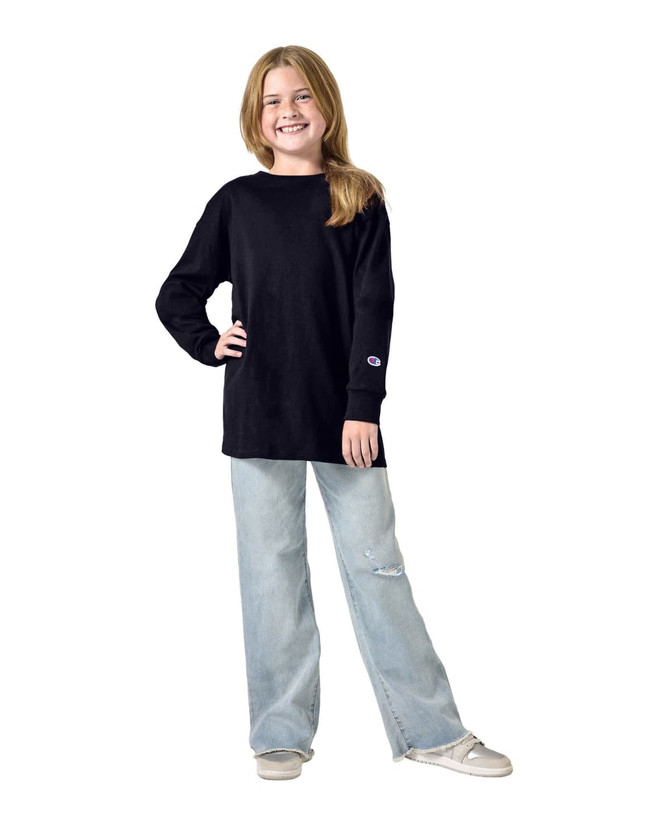 A youth long sleeve T-shirt in black, worn by a smiling girl paired with light blue jeans. The shirt has a logo on the sleeve.