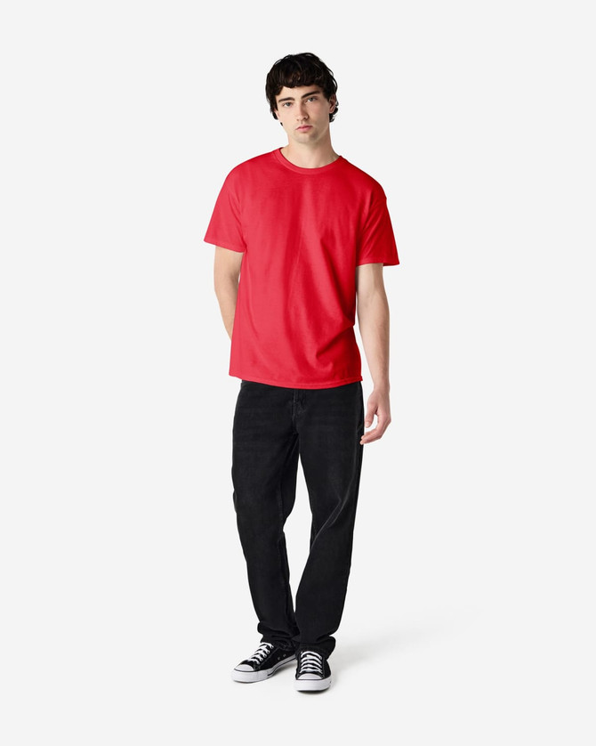 A red Gildan Heavy Cotton Adult T-Shirt worn by a model, paired with black jeans and sneakers. The shirt has a logo.