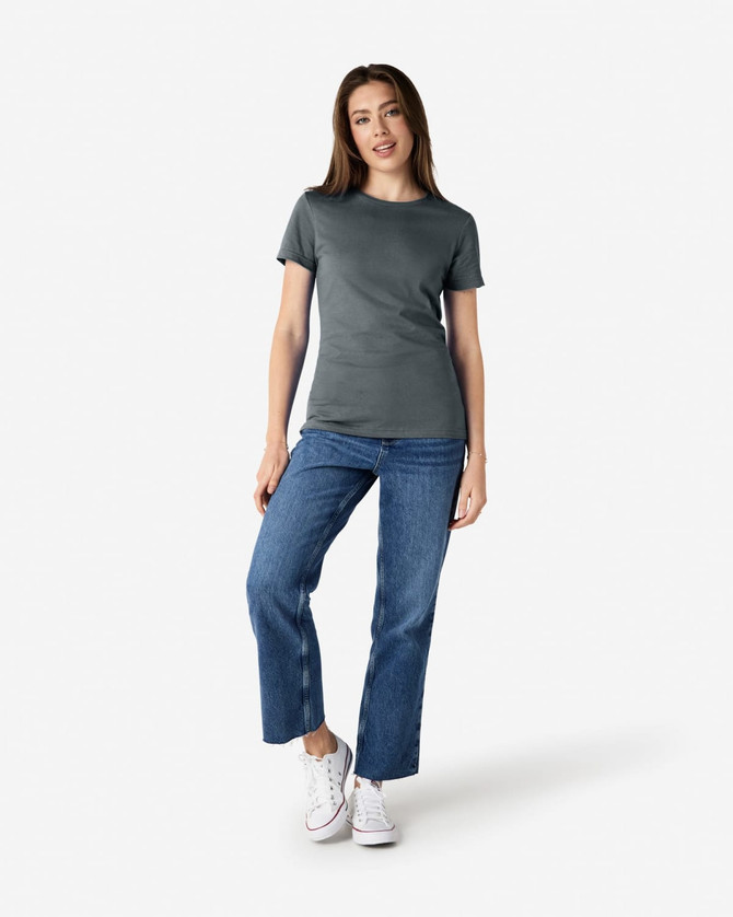 A woman stands wearing a soft-style ladies' t-shirt in grey, paired with blue jeans and white sneakers.