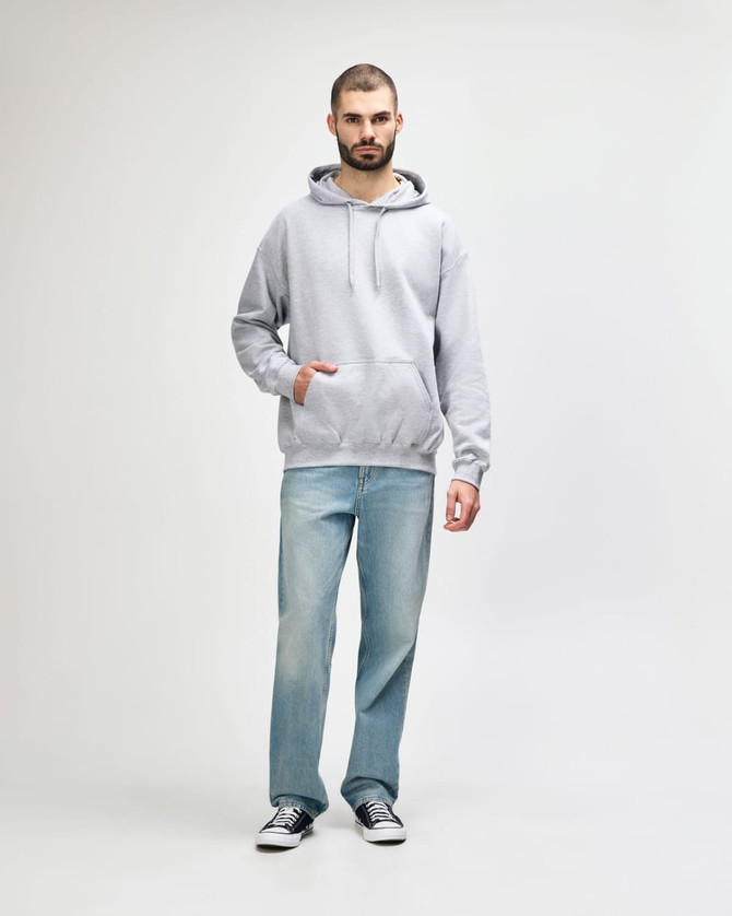 A man stands wearing a grey Gildan Softstyle hooded sweatshirt and light blue jeans, with a logo on the sweatshirt.