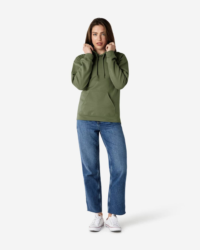 A woman stands wearing a green hooded sweatshirt and blue jeans, with her hands near the hood. The sweatshirt has a logo.