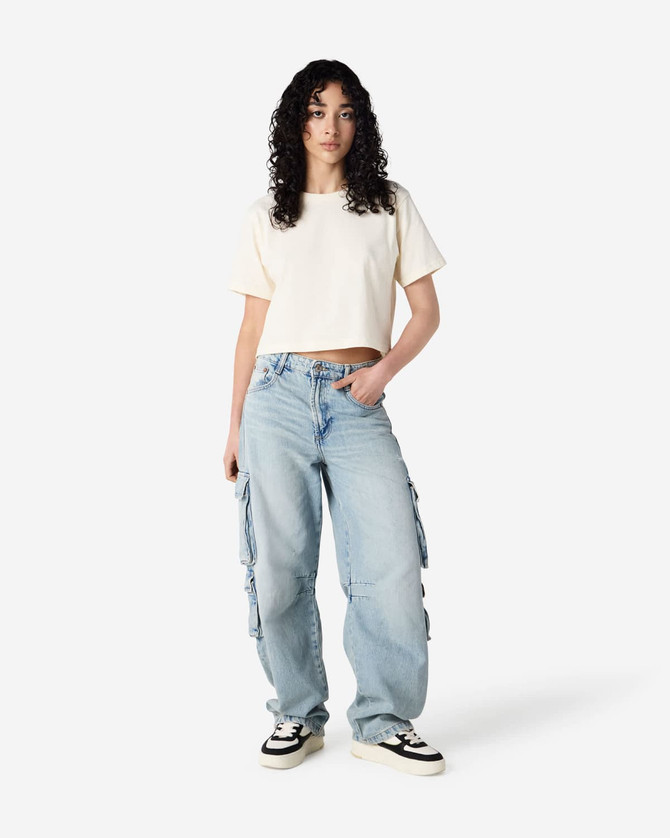 A woman stands in a beige boxy t-shirt and light blue cargo jeans, featuring a logo on the shirt.