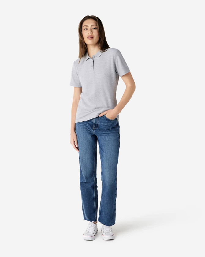 A woman wearing a grey Gildan Softstyle ladies' double pique polo and blue jeans, standing casually.