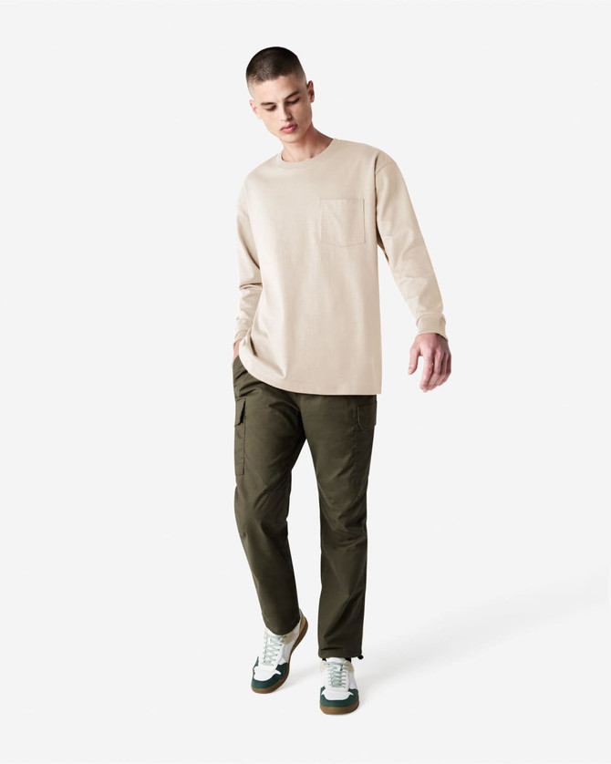 A model wearing a beige long-sleeve T-shirt with a pocket paired with olive green pants and casual sneakers.