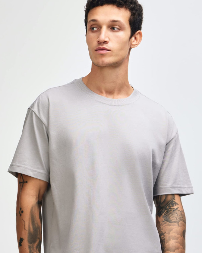 A grey American Apparel Super Heavyweight T-Shirt displayed on a model with curly hair. The shirt has a logo.