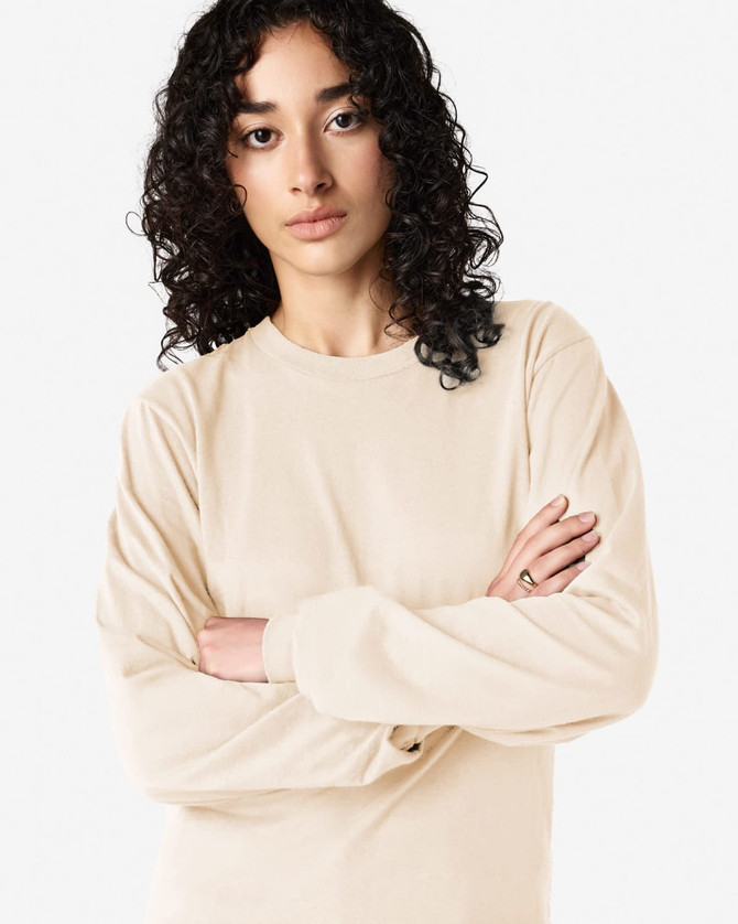 A long sleeve t-shirt in a light beige colour on a model with curly hair, featuring a small logo.