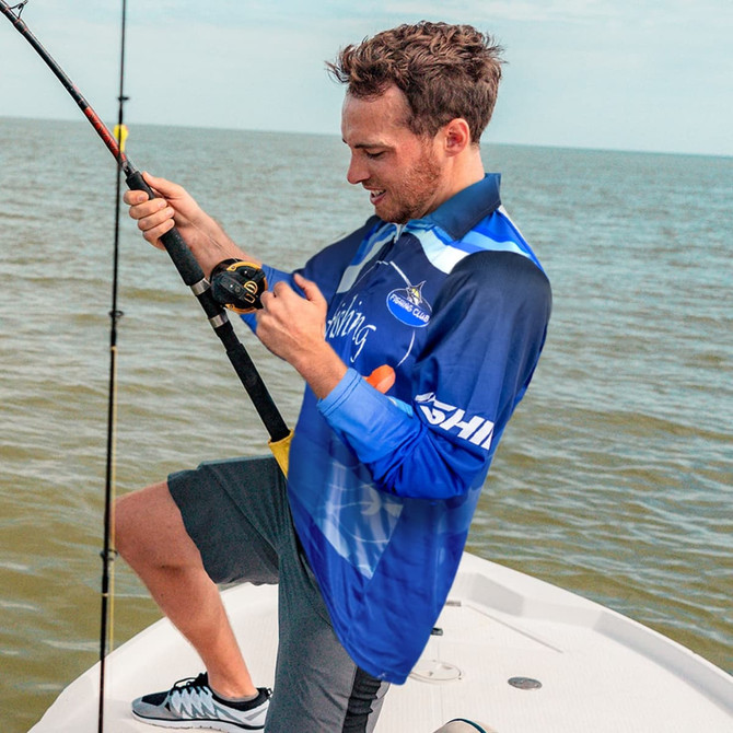 A man wearing a blue custom fishing polo holds a fishing rod on a boat, with water in the background.