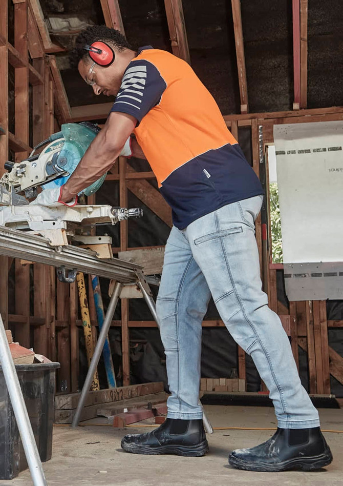 A man wearing an orange and navy short sleeve polo shirt works with a saw in a workshop setting.