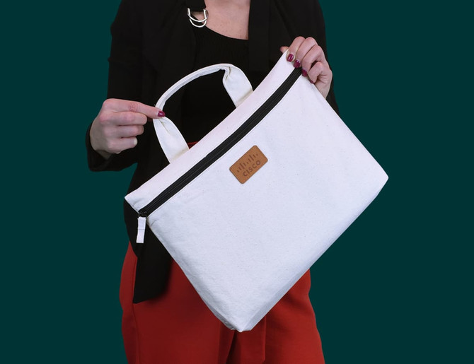 A white conference bag with a black zipper, featuring a logo, held by a person against a dark green background.