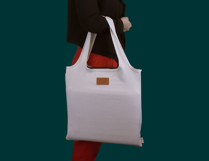 A white tote bag with long handles, featuring a logo, held by a person wearing a black top and red pants.