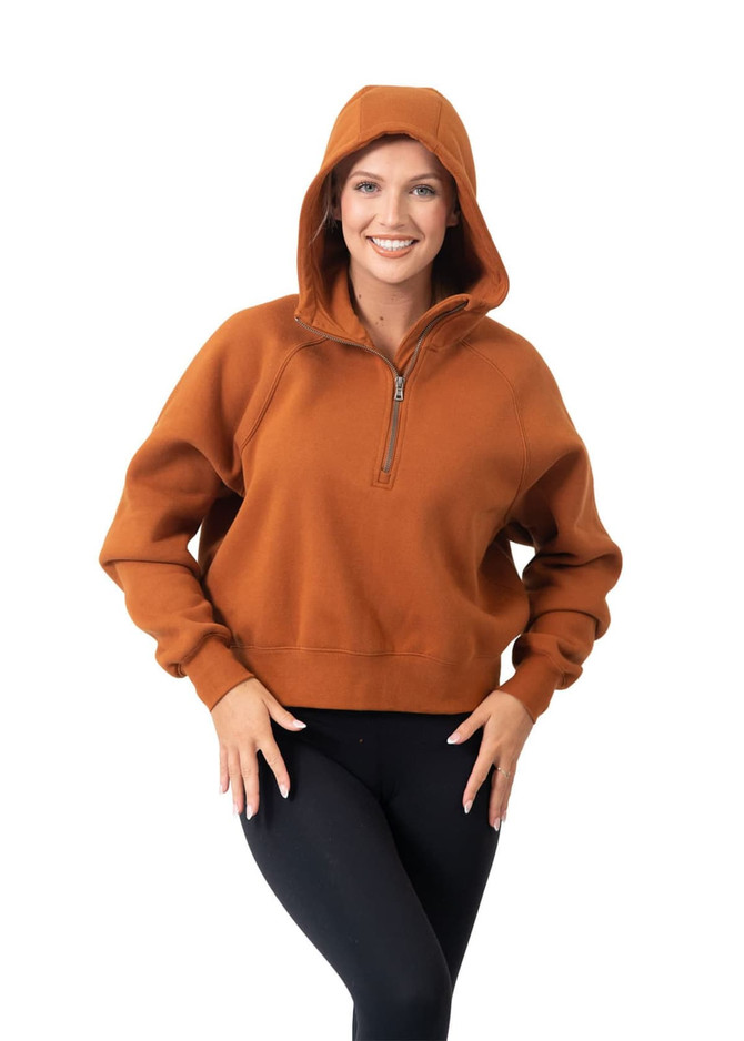 A woman poses in a rust-coloured half-zip hoodie with a hood, paired with black leggings. The hoodie features a small logo.