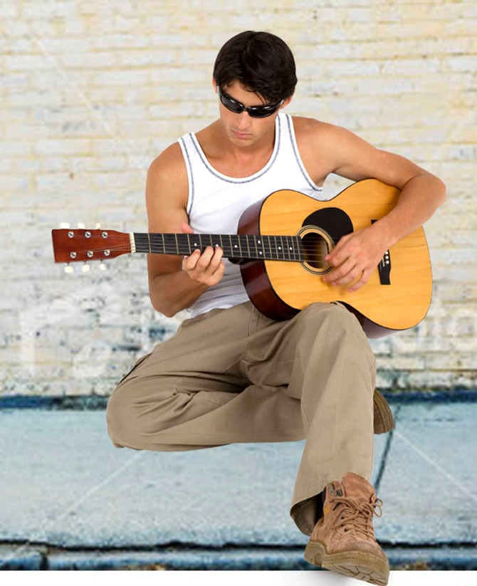 A man wearing a white cotton rib singlet and sunglasses plays an acoustic guitar, sitting against a brick wall.