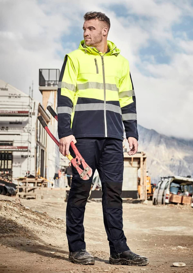 A man in a high-visibility yellow softshell jacket stands on a construction site, holding tools. The jacket features reflective strips.
