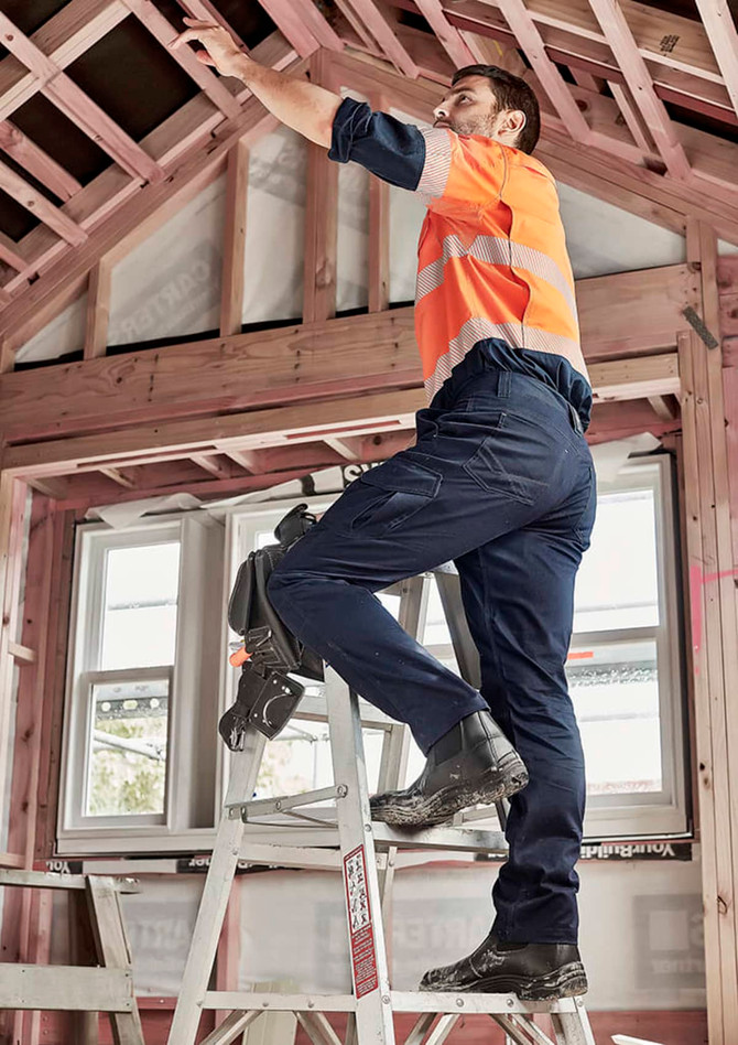 Men's cargo pant in navy, worn by a man on a ladder, working in a partially constructed building. The pants have pockets and a logo.