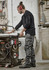 A man wearing black work pants stands by a workshop table, handling tools in a well-equipped space.