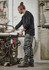 A worker in black Mens Streetworx Heritage Pants, operating machinery in a workshop, with tools and equipment in the background.