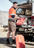 A man in an orange high-visibility shirt and beige shorts stands by an open vehicle boot, holding a tool.