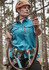 A woman in a blue outdoor short sleeve shirt, wearing a helmet and harness, stands on a zip line in a forest.