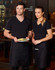 Two individuals wear black short waist aprons over black tops, standing with a plate of food in a restaurant setting.