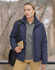 A woman in a navy puffer jacket stands outdoors, holding a book and wearing a grey polo shirt and tan trousers.