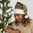A woman wearing a full colour green and white Santa hat, surprised while opening a gift, with a Christmas tree in the background.