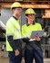 A unisex long-sleeve safety polo in high-visibility yellow with grey segments, featuring two workers wearing hard hats and safety glasses.