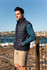 A man stands on rocky beachwearing a navy blue puffer vest and a teal long-sleeve top, with coastline in the background.