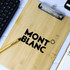 A bamboo clipboard with a dark logo, placed on a surface with a keyboard, pen, and notebook.