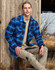 A young man wearing a blue and black quilted flannel shirt poses outdoors, seated against a wooden structure.