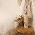 A drink bottle in a soft beige colour with a wooden lid, featuring a logo, sits on a wooden stool beside dried flowers.