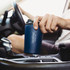 A drink bottle in dark blue, featuring a logo, held in a person's hand inside a vehicle.
