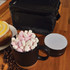 A black coffee mug filled with pink and white marshmallows, next to a silver speaker and a black storage bag.