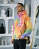 A vibrant tie-dye hoodie featuring shades of pink, orange, yellow, and blue, worn by a model indoors.