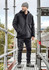 A black unisex softshell jacket worn by a man on scaffolding, with a beanie and work pants, set outdoors.