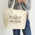 A natural-coloured tote bag with a logo, held by a person wearing a grey sweater and jeans.