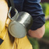 A stainless steel coffee mug with a carabiner attached, featuring a logo, hung from a backpack.