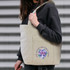 A canvas tote bag in natural colour featuring a floral design and a logo, held by a person wearing a black sweater.