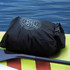 A black lightweight dry bag with a logo, resting on a paddleboard beside calm water.